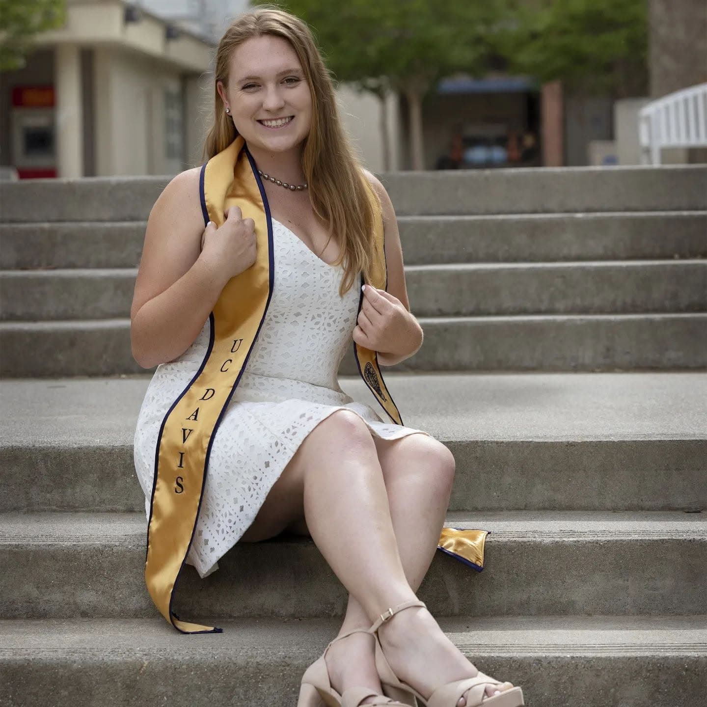 Smiling woman in white lace dress sits on steps wearing a gold UC Davis stole.
