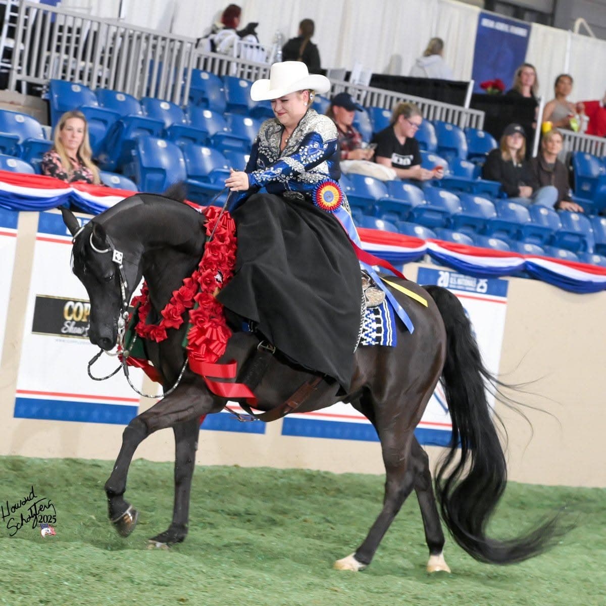 Woman in a white cowboy hat riding a black horse with red rose garland.